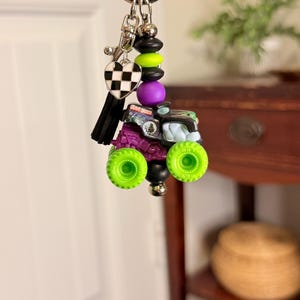 May include: A keychain featuring a miniature monster truck with neon green wheels, purple chassis, and a black and green body. The keychain includes a black and white checkered heart charm, black tassel, and decorative beads.