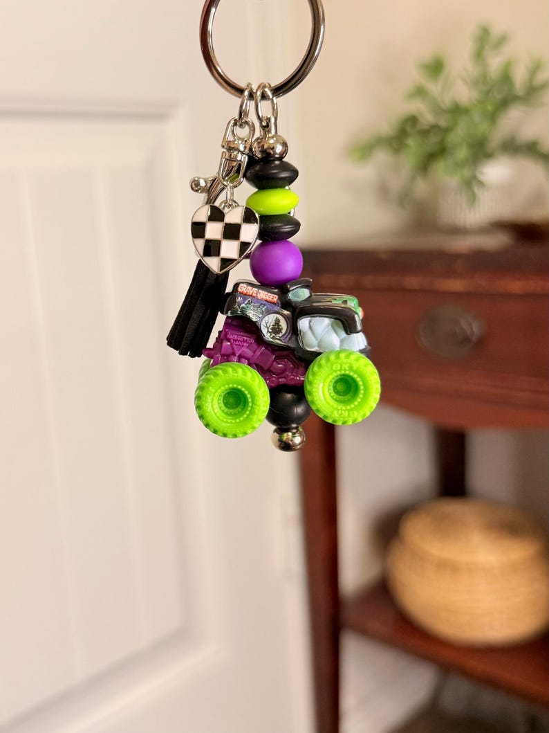May include: A keychain featuring a miniature monster truck with neon green wheels, purple chassis, and a black and green body. The keychain includes a black and white checkered heart charm, black tassel, and decorative beads.