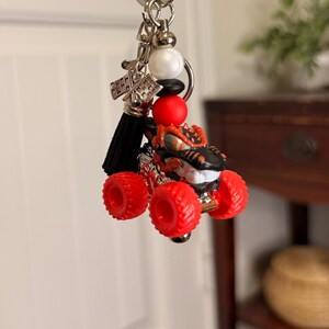 May include: A monster truck keychain with red wheels and a black and orange body. The keychain includes a black tassel, silver accents, and a key ring. The monster truck is a toy.