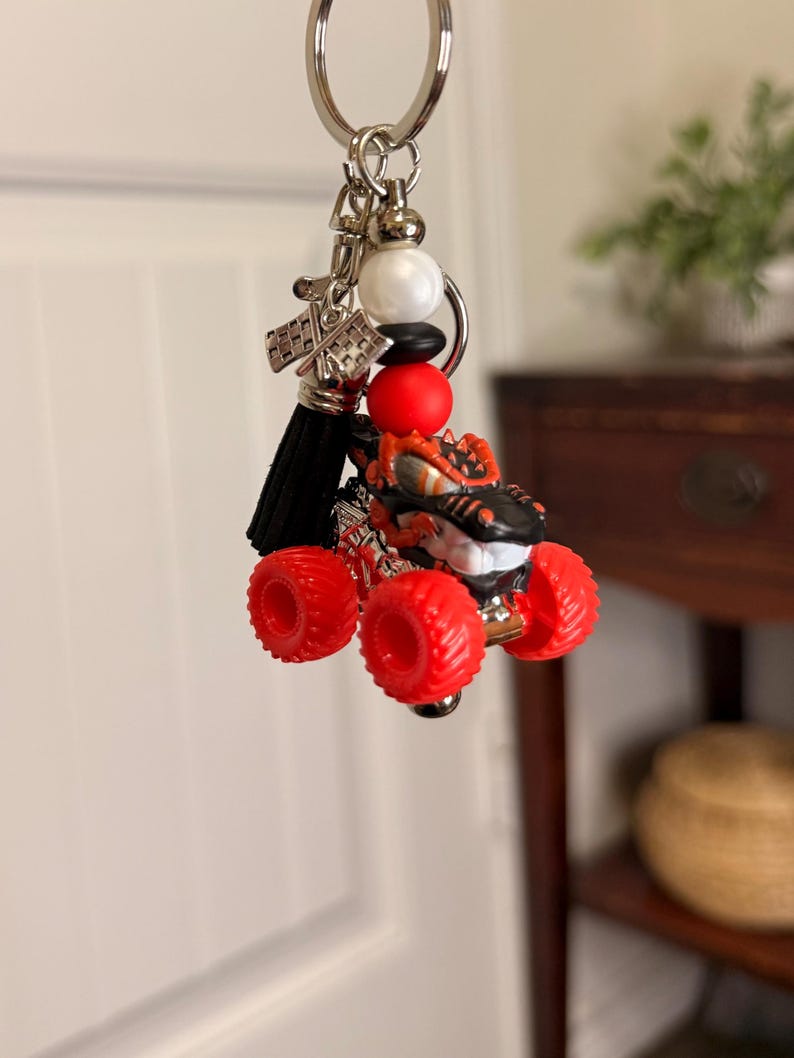 May include: A monster truck keychain with red wheels and a black and orange body. The keychain includes a black tassel, silver accents, and a key ring. The monster truck is a toy.