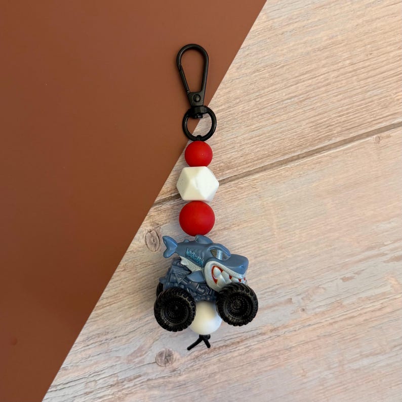 May include: A shark-themed keychain featuring a blue monster truck with black wheels and a cartoon shark design. The keychain includes red and white beads, a black clasp, and a white bead at the bottom. The background is a mix of brown and wood-grain textures.