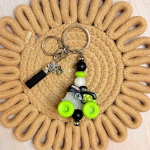 May include: A tractor-themed keychain with a black tassel and a money charm. The tractor charm has green wheels and is accented with black, white, and green beads. The keychain is on a tan woven coaster.