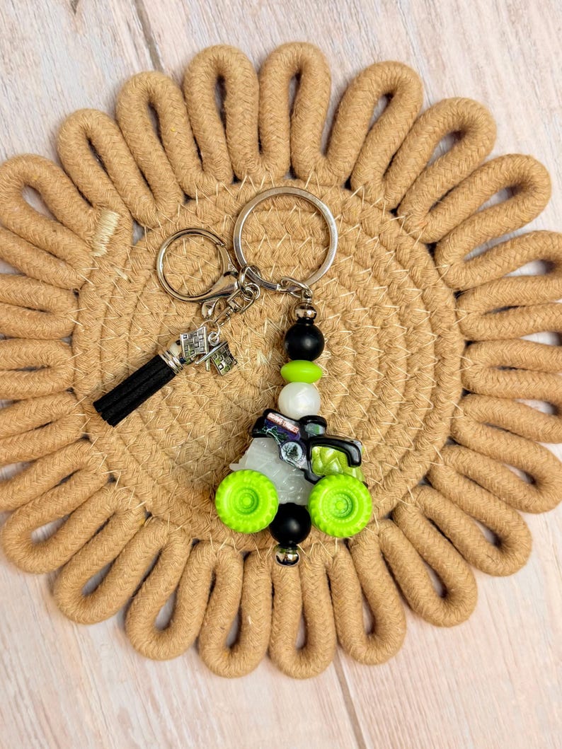 May include: A tractor-themed keychain with a black tassel and a money charm. The tractor charm has green wheels and is accented with black, white, and green beads. The keychain is on a tan woven coaster.