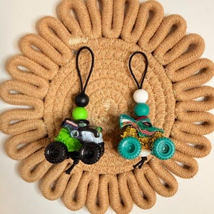 May include: Two toy monster trucks with black wheels and colorful designs. One is green and black, the other is teal and gold. Each truck has a black loop with beads. They rest on a tan, woven, circular mat.
