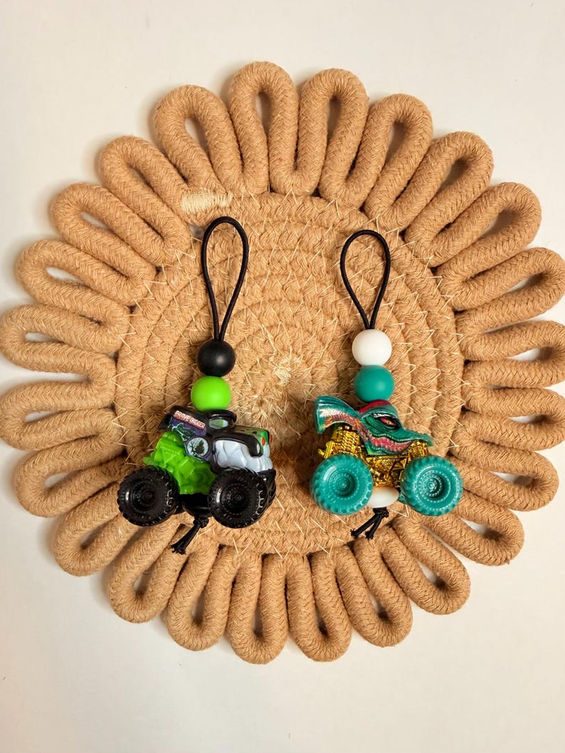 May include: Two toy monster trucks with black wheels and colorful designs. One is green and black, the other is teal and gold. Each truck has a black loop with beads. They rest on a tan, woven, circular mat.