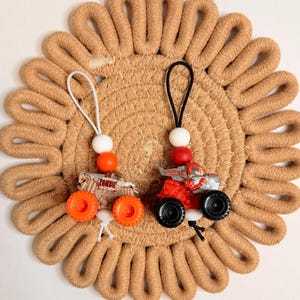 May include: Two miniature monster truck keychains. One truck is red and gray with black wheels, the other is tan with orange wheels and the word "ZOMBIE". Both have a white string with a white bead and a colored bead.