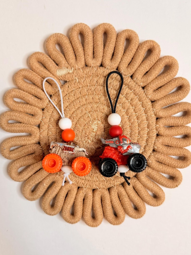 May include: Two miniature monster truck keychains. One truck is red and gray with black wheels, the other is tan with orange wheels and the word "ZOMBIE". Both have a white string with a white bead and a colored bead.