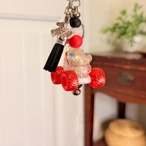 May include: A silver keychain with a clear monster truck charm with red wheels. The keychain also features a black tassel, red and black beads, and a silver charm.