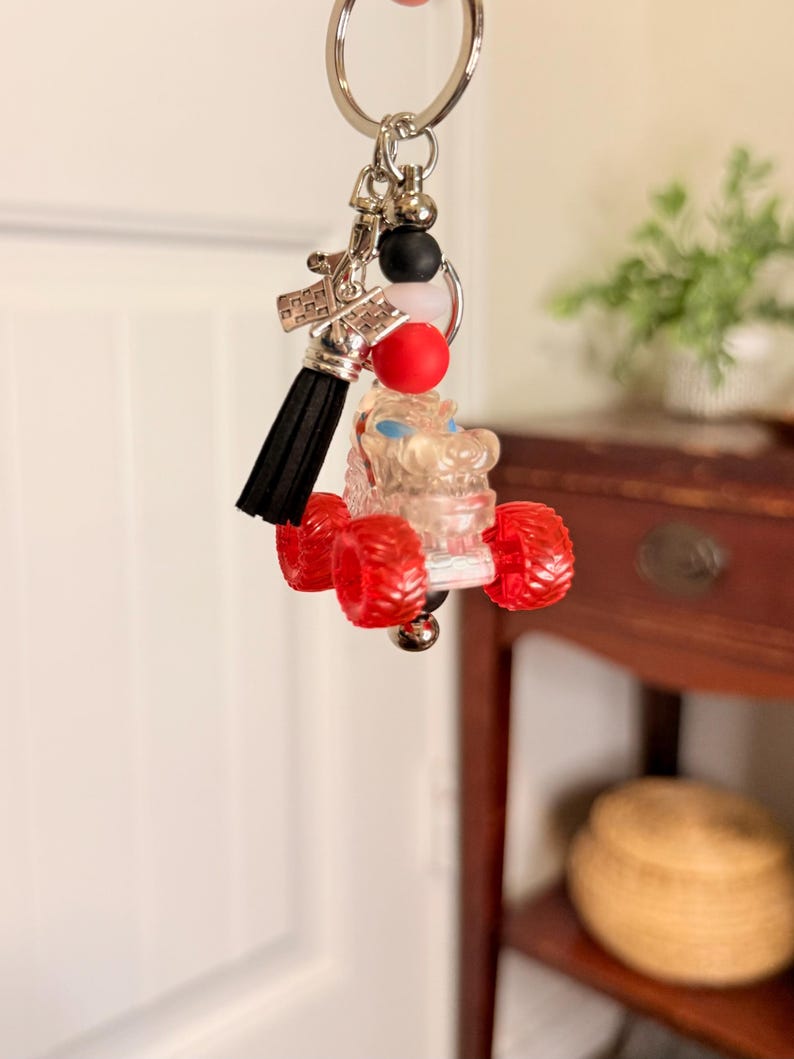 May include: A silver keychain with a clear monster truck charm with red wheels. The keychain also features a black tassel, red and black beads, and a silver charm.