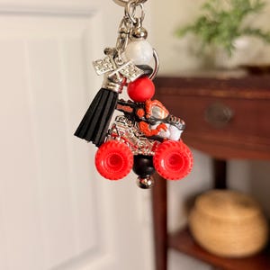 May include: A silver keychain with a miniature monster truck charm. The truck features red wheels, a black body, and orange and silver accents. A black tassel and decorative beads complete the keychain.