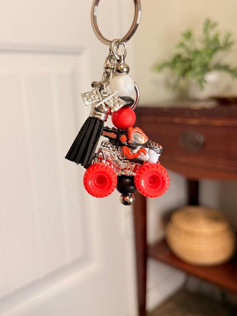May include: A silver keychain with a miniature monster truck charm. The truck features red wheels, a black body, and orange and silver accents. A black tassel and decorative beads complete the keychain.