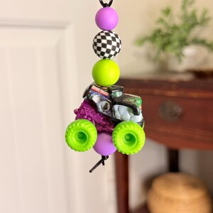 Pu&ograve; includere: Un portachiavi nero con una serie di perline colorate e un ciondolo a forma di monster truck in miniatura. Le perline includono una sfera viola, una palla a scacchi in bianco e nero e una sfera verde brillante. Il monster truck ha ruote verdi.