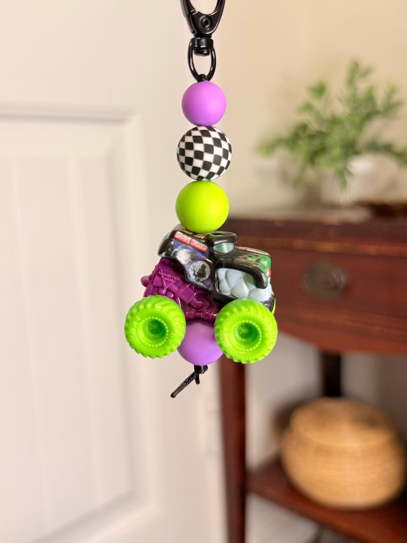 Pu&ograve; includere: Un portachiavi nero con una serie di perline colorate e un ciondolo a forma di monster truck in miniatura. Le perline includono una sfera viola, una palla a scacchi in bianco e nero e una sfera verde brillante. Il monster truck ha ruote verdi.