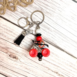May include: A silver keychain with a black tassel and a miniature race flag charm. The keychain features a red and black ATV charm with red wheels and a black bead. Additional beads are white, black, and red.