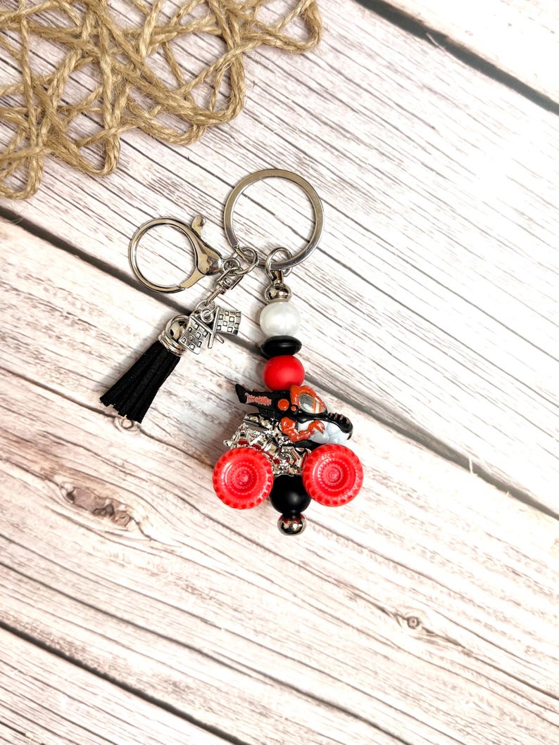 May include: A silver keychain with a black tassel and a miniature race flag charm. The keychain features a red and black ATV charm with red wheels and a black bead. Additional beads are white, black, and red.