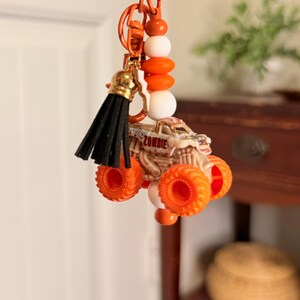 May include: Orange monster truck keyring with the word "ZOMBIE" on the side. The keyring has a black tassel and orange and white beads. The keyring is orange.