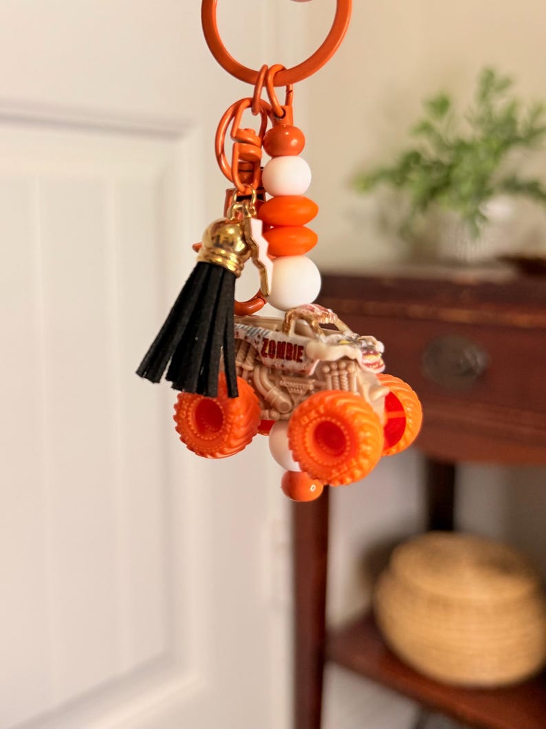 May include: Orange monster truck keyring with the word "ZOMBIE" on the side. The keyring has a black tassel and orange and white beads. The keyring is orange.