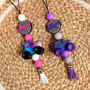 May include: Two beaded lanyards with a black cord. Each features a 'Mama' charm, a monster truck, and a tassel. One lanyard has a blue truck, the other a purple truck. The charms are accented with pink, white, and black beads.