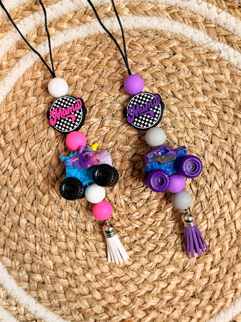 Op de afbeelding: Twee kralen lanyards met een zwart koord. Elk heeft een 'Mama' bedel, een monstertruck en een kwastje. E&eacute;n lanyard heeft een blauwe truck, de andere een paarse truck. De bedels zijn geaccentueerd met roze, witte en zwarte kralen.