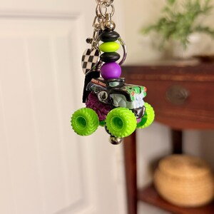 May include: A keychain featuring a miniature monster truck toy with neon green wheels and a black and green body. The keychain includes a charm with the words "Grave Digger" and a checkered flag charm, along with decorative beads in black, green, and purple.