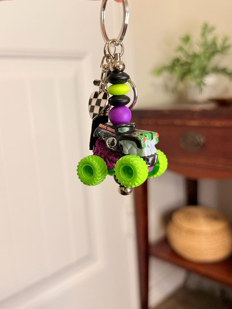 May include: A keychain featuring a miniature monster truck toy with neon green wheels and a black and green body. The keychain includes a charm with the words "Grave Digger" and a checkered flag charm, along with decorative beads in black, green, and purple.