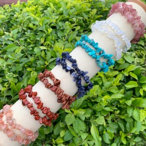 Può includere: Una collezione di braccialetti con schegge di pietre preziose in vari colori, tra cui rosa, bianco, turchese, blu e rosso. I braccialetti sono esposti su una forma cilindrica bianca su uno sfondo di fogliame verde. Ogni braccialetto è fatto di piccole pietre di forma irregolare.
