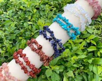 Puede incluir: Una colección de pulseras de virutas de piedras preciosas en varios colores, incluyendo rosa, blanco, turquesa, azul y rojo. Las pulseras se exhiben en una forma cilíndrica blanca sobre un fondo de follaje verde. Cada pulsera está hecha de piedras pequeñas de forma irregular.