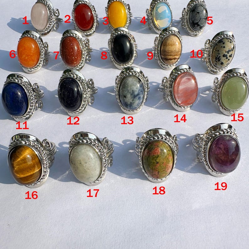 May include: A collection of nineteen silver-toned rings, each featuring a different oval-shaped gemstone. The gemstones vary in color, including pink, red, yellow, black, and green. The rings are displayed in a grid, showcasing the variety of designs.