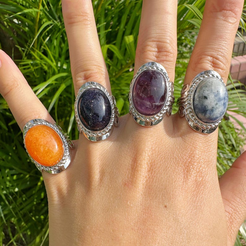 May include: Four gemstone rings with oval-shaped stones in orange, dark blue, purple, and blue-gray hues. Each ring has a silver-colored, textured band. The rings are displayed on a hand against a backdrop of green foliage.