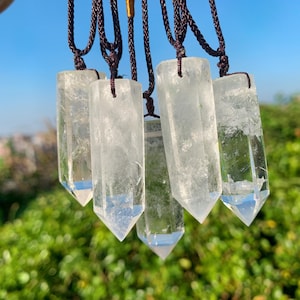 Può includere: Ciondoli di cristallo di quarzo trasparente appesi a cordoncini marroni. Ogni cristallo è un prisma rettangolare con una punta. I cristalli sono traslucidi con lievi inclusioni interne. Lo sfondo è un fogliame verde sfocato.