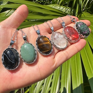 May include: A selection of gemstone pendant necklaces. Each necklace has an oval-shaped stone in a silver setting, with a chain. The stones vary in colour and pattern, including black and white, green, brown, clear, red, and green with red.