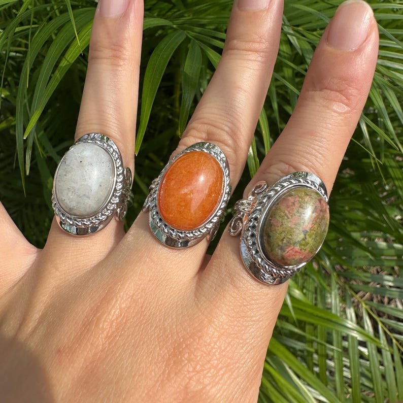 May include: Three gemstone rings with oval-shaped stones in white, orange, and green-brown hues. The rings have silver-colored settings with decorative rope-like and swirl details. The rings are displayed on a hand against a backdrop of green foliage.