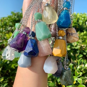 May include: A selection of gemstone pendant necklaces in a variety of colours, including purple, blue, green, yellow, and clear. Each pendant is a faceted, teardrop-shaped bottle with a silver chain. The pendants are held against a backdrop of green foliage.