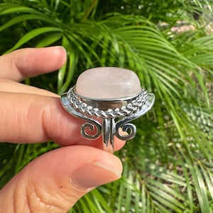 May include: A silver ring featuring a large, oval rose quartz gemstone. The gemstone is a soft pink color and is set in a decorative silver setting with a braided design around the stone. The ring has an ornate, swirling design on the sides.