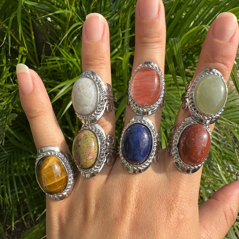 May include: A hand displays seven gemstone rings. Each ring features a different oval-shaped stone in colors like yellow, green, red, blue, and white, set in a silver-colored metal band with a decorative edge.