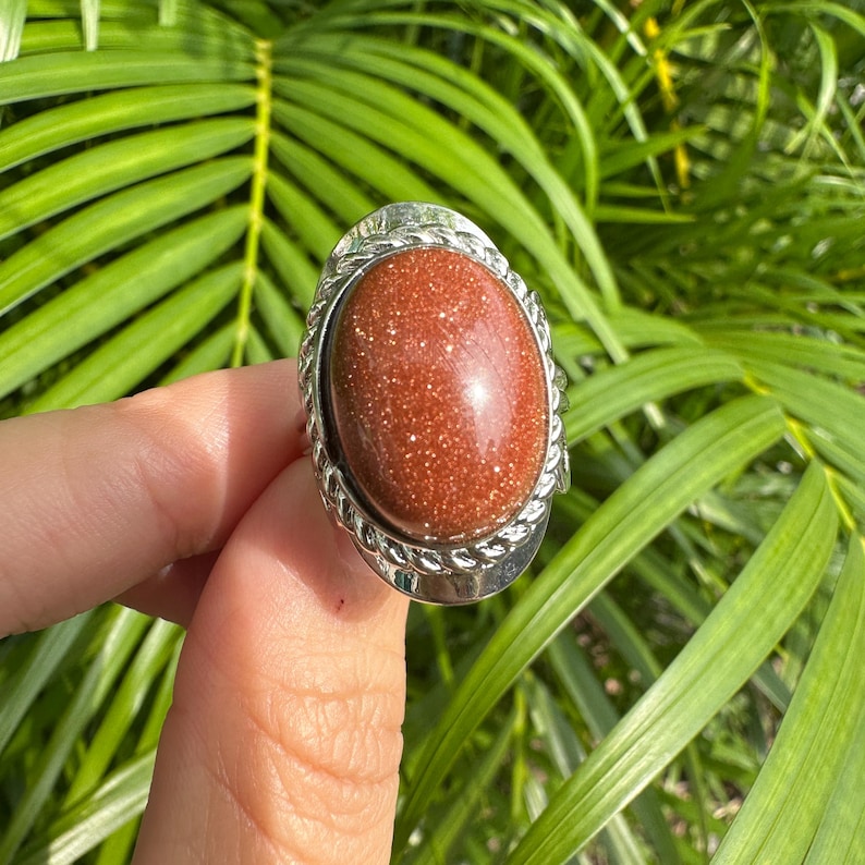 May include: Oval-shaped ring featuring a large, sparkling, reddish-brown stone set in a silver-colored metal band. The band has a decorative rope-like design around the stone. The ring is displayed against a backdrop of green foliage.