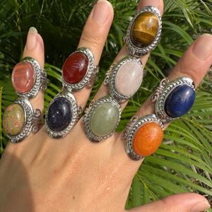 May include: A hand displays a collection of gemstone rings. Each ring features a different oval-shaped stone in colors like red, blue, orange, and green, set in a silver-toned metal band with a decorative rope-like border.
