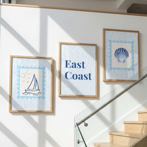 Op de afbeelding: Drie ingelijste wandposters met lichtblauwe verticale strepen. De linker poster toont een zeilboot, de middelste poster bevat de tekst East Coast en de rechter poster toont een blauwe schelp. Elke poster heeft een lichtblauwe postzegelrand.