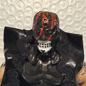 May include: A black figurine of a masked wrestler with a red and black design on the mask. The wrestler wears a black outfit with a cape trimmed with a black, wavy design. The figure is standing on a silver base.