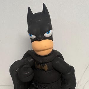 May include: A black Batman figurine with a tan face, blue eyes, and a bat symbol on the chest. The figurine has a cape and is posed with one arm forward. The figure is standing on a white surface.