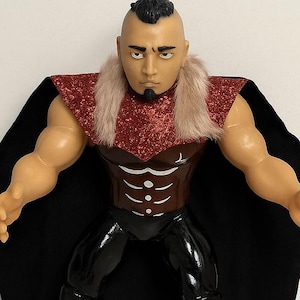 Puede incluir: Una figura de acción artesanal de 18 cm de El Hijo Del Vikingo, un luchador de Lucha Libre. La figura tiene una constitución musculosa, una capa negra y botas blancas. La figura está sobre una base extraíble y es un objeto de colección de lucha libre mexicana.