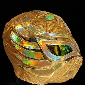 May include: A gold lucha libre mask with iridescent accents. The mask features a mesh design with gold sequins and decorative shapes. The eye and mouth openings are outlined with iridescent material, creating a striking visual effect. The mask is displayed against a black background.
