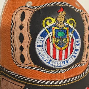 May include: An orange and black baseball cap featuring a central patch with the Club Deportivo Guadalajara crest. The patch is surrounded by tan leather detailing with cutouts, black and white braided trim, and a black leather-like base panel.