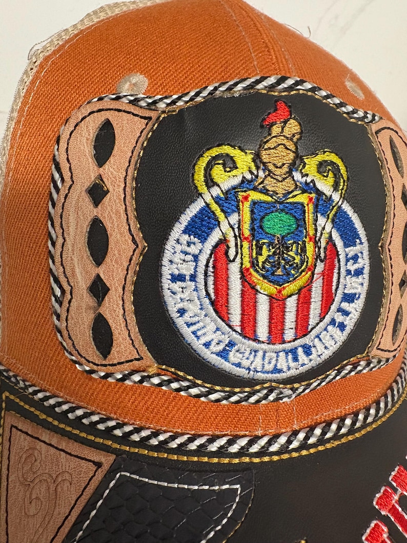 May include: An orange and black baseball cap featuring a central patch with the Club Deportivo Guadalajara crest. The patch is surrounded by tan leather detailing with cutouts, black and white braided trim, and a black leather-like base panel.