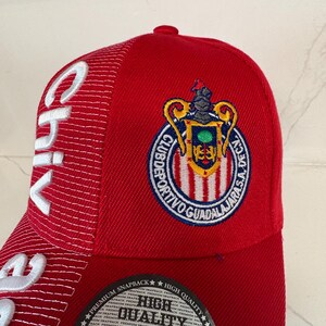 May include: A red snapback cap featuring the Club Deportivo Guadalajara crest embroidered in blue, yellow, and red. The side panel displays white embroidered text reading Chivas, and a circular black sticker on the brim reads High Quality.
