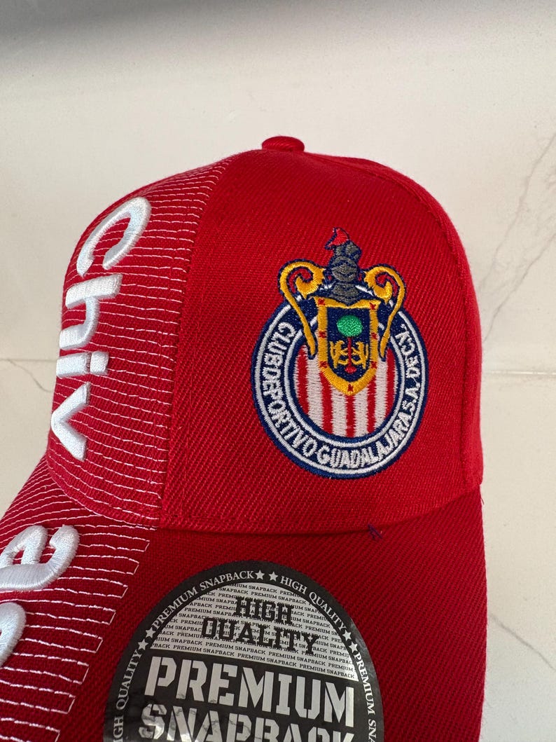 May include: A red snapback cap featuring the Club Deportivo Guadalajara crest embroidered in blue, yellow, and red. The side panel displays white embroidered text reading Chivas, and a circular black sticker on the brim reads High Quality.