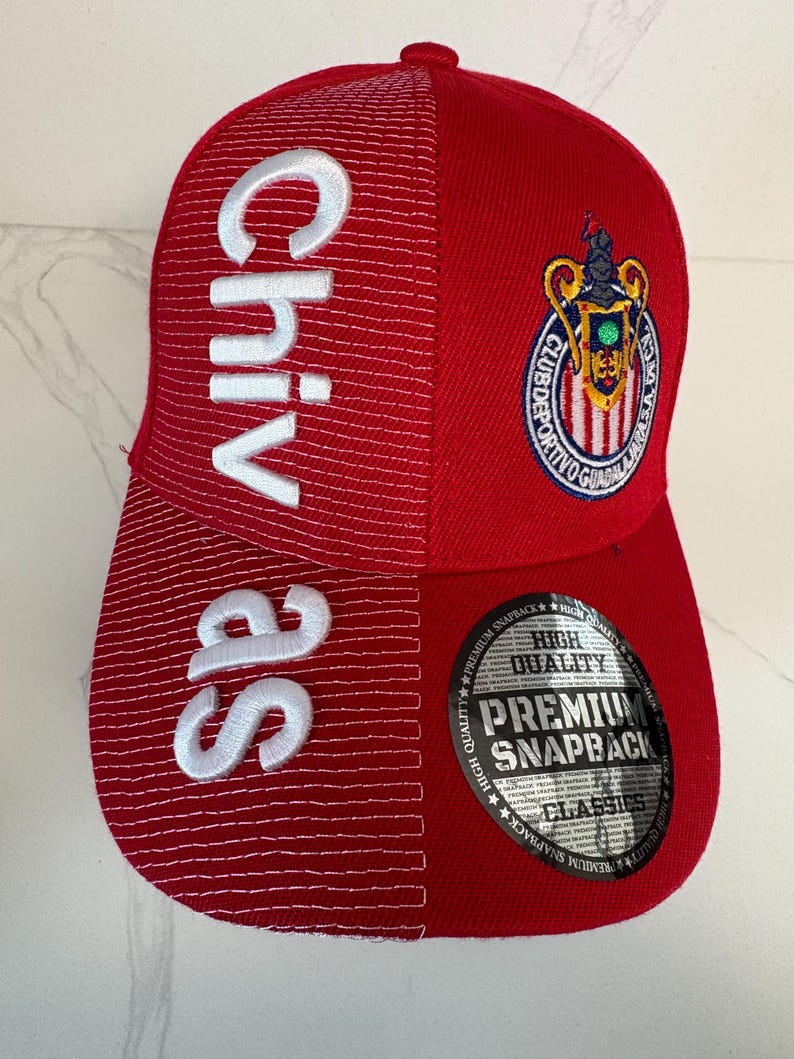 May include: A red snapback cap featuring white embroidered text reading Chivas on the left side and a colorful Club Deportivo Guadalajara crest on the right. A circular black and silver sticker on the brim displays the text High Quality Premium Snapback Classics.