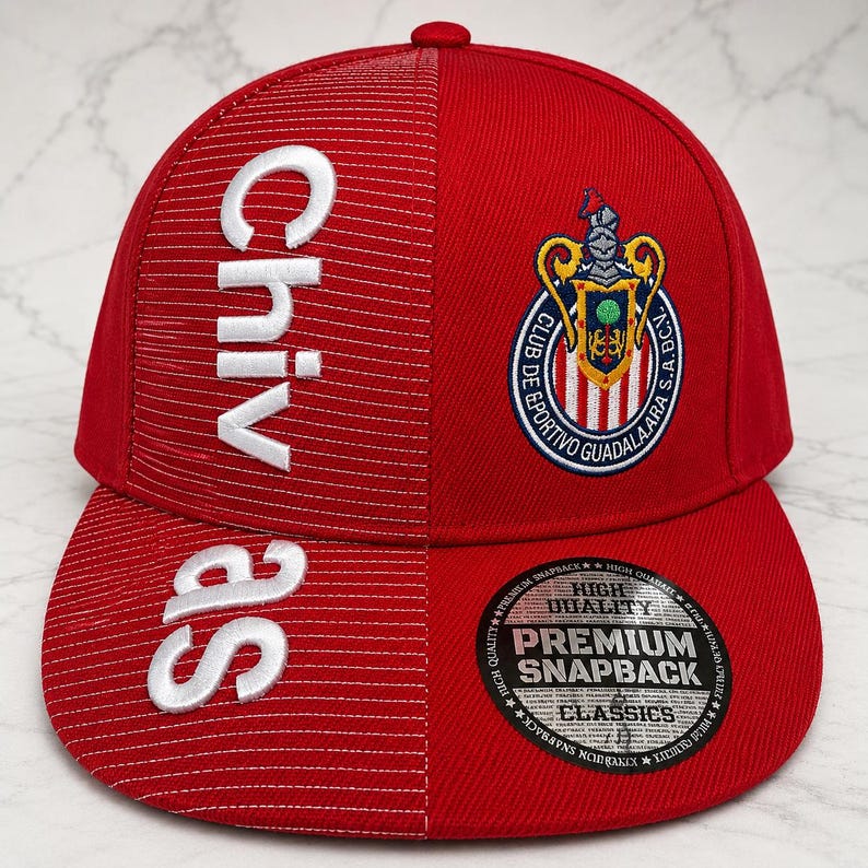 May include: A red snapback cap featuring a textured left panel with the white embroidered text Chivas. The right panel displays the embroidered Club Deportivo Guadalajara crest. A circular black and silver sticker is on the flat brim.