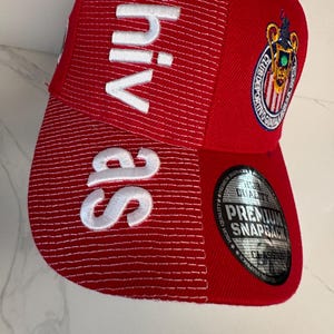 May include: A red snapback cap featuring white embroidered text that reads Chivas vertically on the front panel. The cap includes a circular embroidered patch of the Club Deportivo Guadalajara logo and a round silver sticker on the brim.