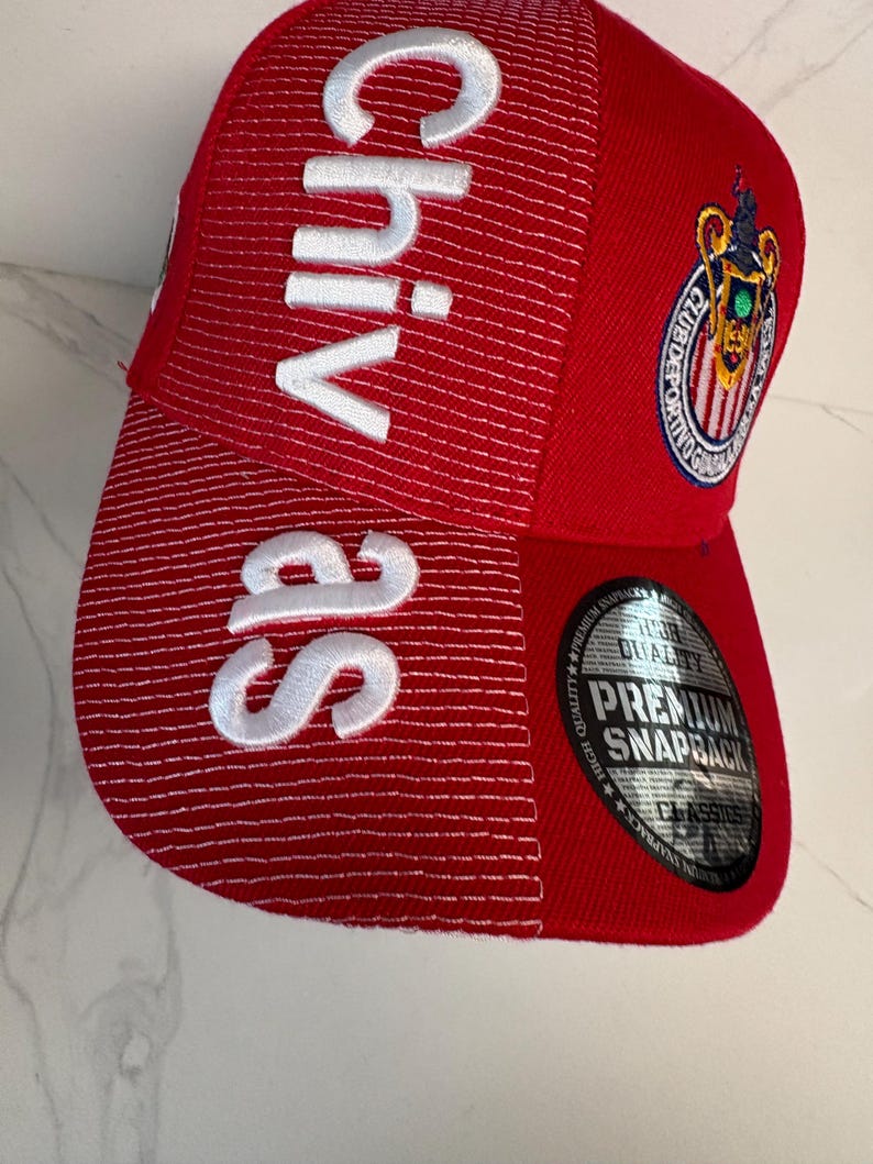 May include: A red snapback cap featuring white embroidered text that reads Chivas vertically on the front panel. The cap includes a circular embroidered patch of the Club Deportivo Guadalajara logo and a round silver sticker on the brim.